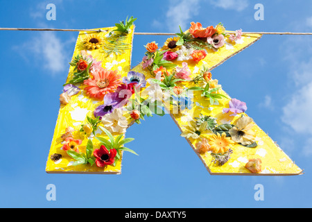 Cardboard cutout of the letter K, painted yellow and covered in flowers. Hanging on a line against a blue summer sky. Stock Photo
