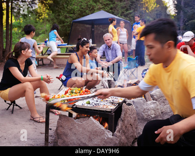 Group of people of different ethnicities enjoying themselves sitting around a fire at a camp site in a park. Ontario, Canada. Stock Photo