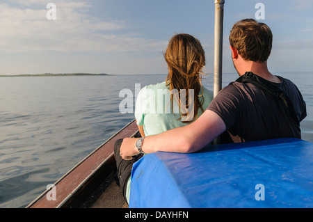A young couple sit at the bow of a boat while approaching an island Stock Photo