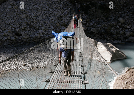 Porters carrying heavy load over a metal suspension bridge in Annapurna Conservation Area, Annapurna Circuit, Nepal Stock Photo