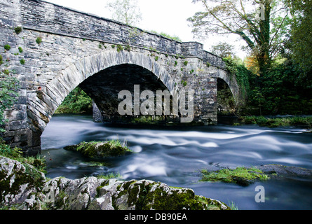 A long exposure shot of a stone bridge, with smooth river water running under and few rocks in the foreground. Skelwith bridge. Stock Photo