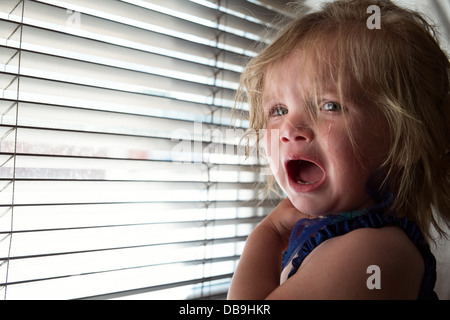 A blond female Caucasian infant crying & distressed by a window covered with venetian blinds Stock Photo