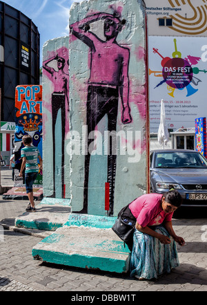 Woman sits at base of painted fragment of Berlin wall at Checkpoint Charlie Border post, Friederichstrasse, Berlin Stock Photo