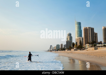 Surfer walking into surf with city skyline in background. Surfers Paradise, Gold Coast, Queensland, Australia Stock Photo