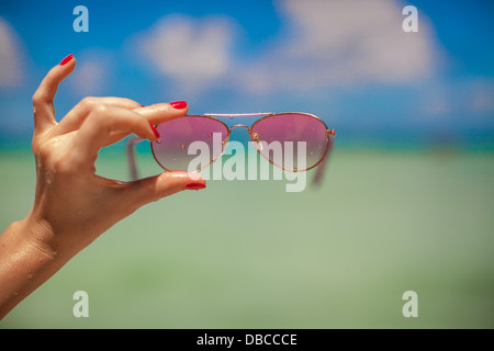 Woman's hand holding pink sunglasses on tropical beach background the sea Stock Photo