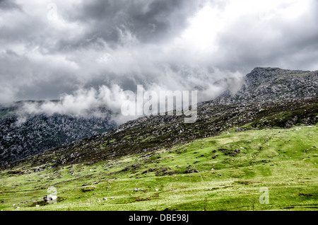 Snowdonia National Park Mountains Wales A4086 Road | SNOWDONIA NATIONAL PARK, Wales — Storm clouds cling to the rocky ridgeline above a green valley along the A4086, a scenic route through the Ogwen and Nant Peris area of Eryri. The mountains here are built from Ordovician volcanic rock roughly 450 million years old, later shaped by glaciers into the steep, boulder-strewn slopes visible in this frame. Stock Photo