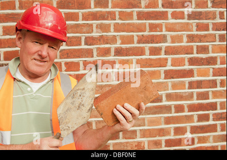 Bricklayer wearing a red safety helmet, holding a brick and trowel in both hands. Stock Photo