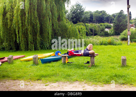 A young male canoeist tries to rest lying on a flat short wooden bench, covering his head with shirt near the river Severn Stock Photo