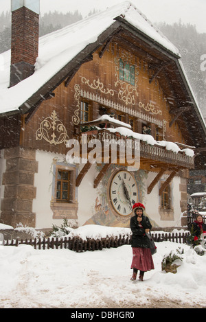 Germany, Black Forest, Hollsteig. Hofgut Sternen Village. Woman in 'pompom' hat in front of cuckoo clock on building. Stock Photo