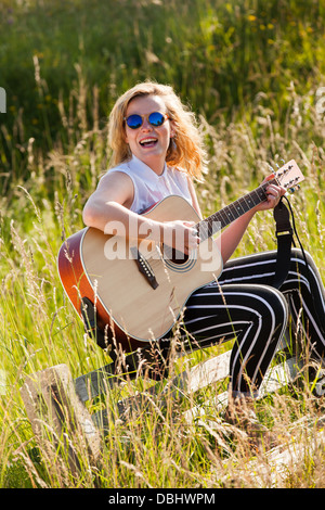 Teenage girl wearing dark glasses playing guitar in a grassy field. Outdoors. Summer. Stock Photo