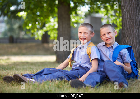 Image of happy twin boys with backpacks sitting in summer park Stock Photo