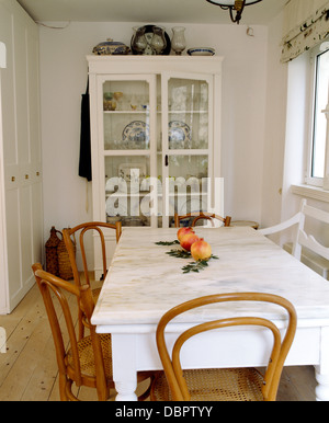 Marble-topped painted table with antique Bentwood chairs in white coastal dining room with white glass-front cupboard Stock Photo