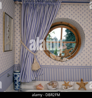 Circular window with blue+white striped curtain in cottage bathroom with blue+white wallpaper and striped wallpaper border Stock Photo