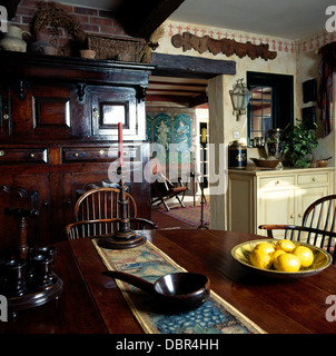 Cotton runner and bowl of grapefruit on antique table with stick-back chairs in cottage dining room with large antique cupboard Stock Photo