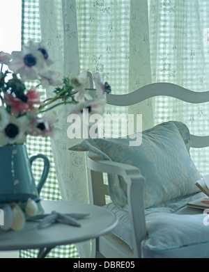Vase of anemones on table beside white chair with white cushions in front of window with floral and checked curtains Stock Photo