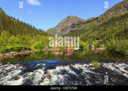 View across flowing water to typical Norwegian farm house and red barn reflected in Smorkleppai River in summer. Grungedal Telemark Norway Stock Photo