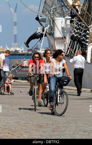 Young, female tourists cycling along the quay in the port of Copenhagen, Denmark. The tall ship DANMARK moored in the background. Stock Photo