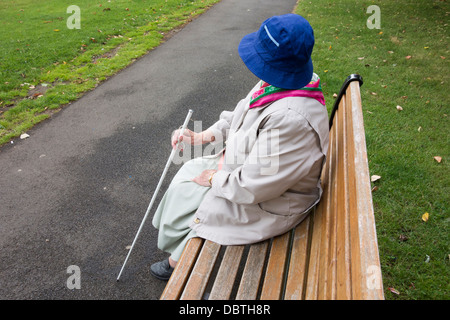 Ninety year old visually impaired lady with white stick sitting on bench in public park. England, UK Stock Photo