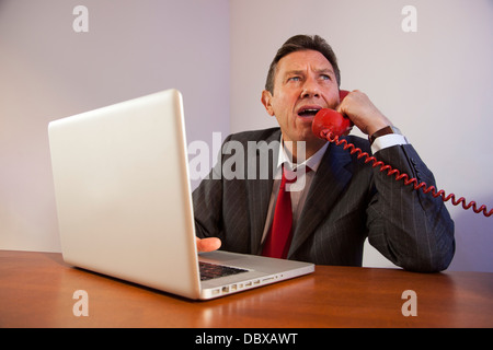 Angry man wearing a suit, shouting down a red telephone while sitting in front of a laptop on a desk. Stock Photo