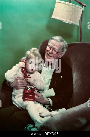 1940s 1950s GRANDDAUGHTER SITTING ON SMILING GRANDFATHER’S LAP TALKING ON TOY TELEPHONE Stock Photo