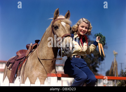 1940s 1950s SMILING YOUNG BLONDE COWGIRL SITTING ON FENCE POSING BY PALOMINO HORSE HOLDING BUNCH OF CARROTS LOOKING AT CAMERA Stock Photo