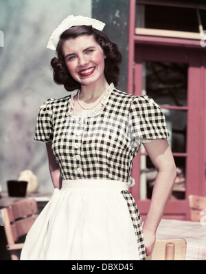 1940s 1950s SMILING WOMAN WEARING BLACK AND WHITE CHECKED WAITRESS OUTFIT DRESS APRON LACE CAP LOOKING AT CAMERA Stock Photo