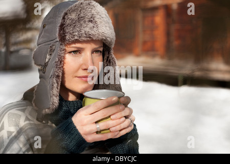 Pensive woman in fur hat drinking coffee outside cabin Stock Photo