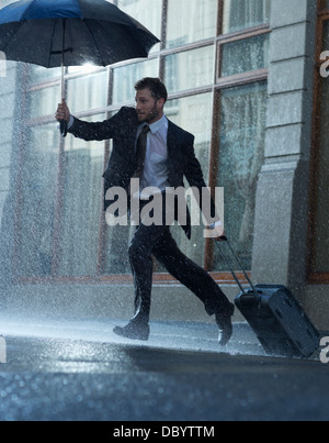 Businessman with suitcase and umbrella crossing rainy street Stock Photo