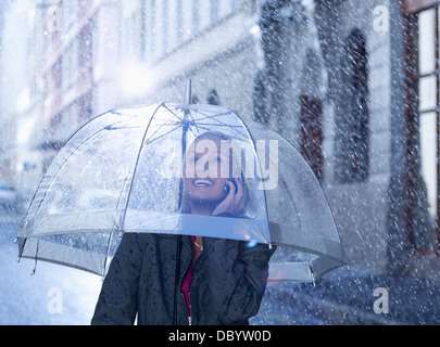 Smiling businesswoman talking on cell phone under umbrella in rainy street Stock Photo