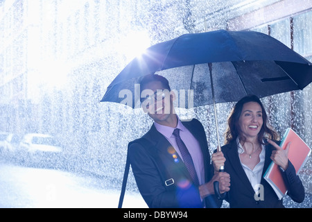 Happy businessman and businesswoman walking under umbrella in rain Stock Photo