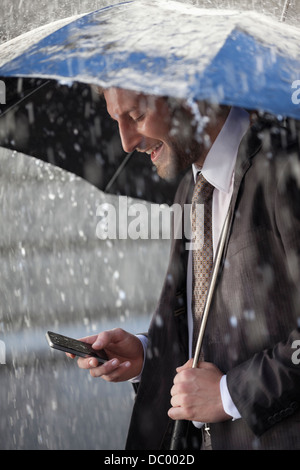 Businessman text messaging on cell phone under umbrella in rain Stock Photo
