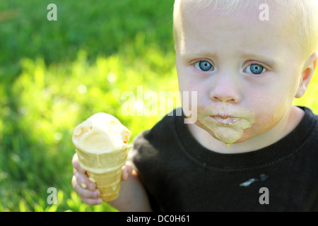 a cute baby boy is sitting outside in the grass on a summer day, eating a messy, melting vanilla ice cream cone. Stock Photo
