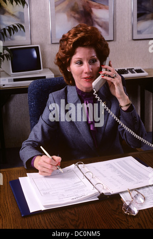 1980s BUSINESSWOMAN TALKING ON TELEPHONE WRITING IN BINDER Stock Photo