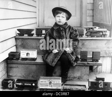 1880s 1890s 1900s BOY WEARING FUR COAT SITTING ON STEPS SURROUNDED BY TOY RAILROAD TRAINS LOOKING AT CAMERA Stock Photo