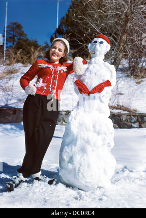 1940s 1950s SMILING TEEN GIRL POSING LEANING AGAINST SNOWMAN LOOKING AT CAMERA LOOKING AT CAMERA Stock Photo