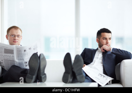 A young serious businessman with newspaper looking at camera with his pensive colleague sitting near by Stock Photo