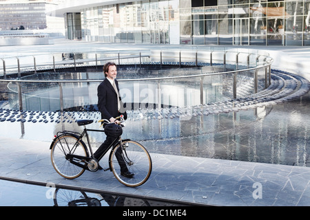 Mid adult businessman walking with bicycle by water feature in city Stock Photo