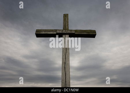 A cross made from English oak rises up into threatening grey English skies. With splits in its vertical and horizontal beams, the word Peace is written in lettering in the centre in an image of Christian values - a message for mankind, of humanity and goodwill to all Men. The wood is from the royal estate of Sandringham in Norfolk, presented in August 1987 by Queen Elizabeth II to mark the site of the high altar of the former St Benet's Abbey near Ludham on the Norfolk Broads. Stock Photo