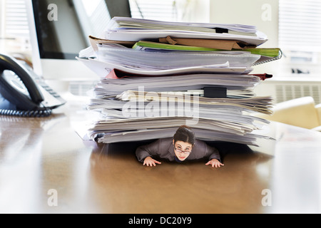 Businesswoman trapped underneath stack of large documents on oversized desk, portrait Stock Photo