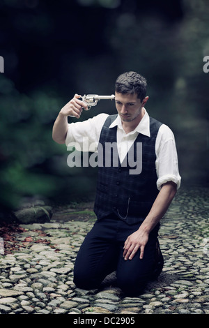 a man in vintage clothes is sitting on the ground, trying to shoot himself Stock Photo