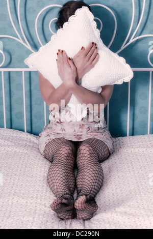 a woman in a negligee is sitting on a bed, a pillow in front of her face Stock Photo