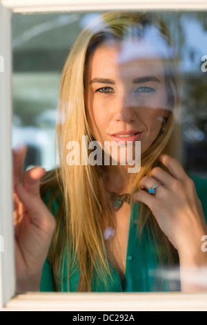 Portrait of young woman looking through window, hand in hair Stock Photo