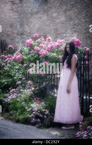 a beautiful woman with long black hair standing in a vintage pink dress in front of a gate with flowers Stock Photo