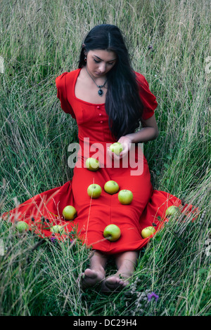 a woman in a red dress sitting on a field, with green apples on her dress Stock Photo
