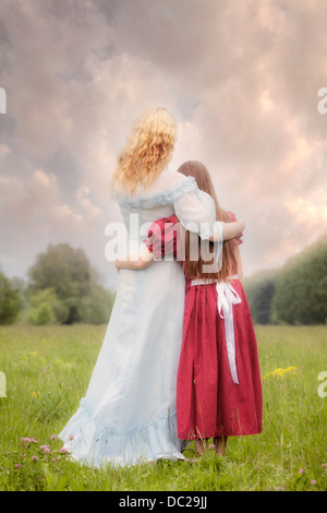 two girls in vintage dresses standing on a meadow, embracing each other Stock Photo