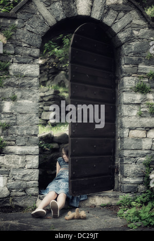 a sad, young girl is sitting in an old door with a teddy bear Stock Photo