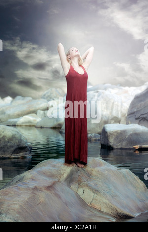 a woman in a red dress is standing on a rock in the water and enjoying the sun Stock Photo