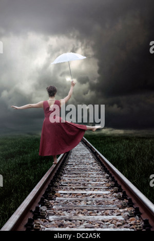 a girl in a red dress is balancing on railway tracks with a white parasol Stock Photo