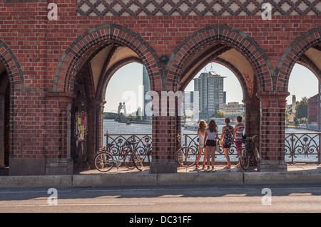 Young women with bicycles standing on the Oberbaum Bridge looking at the Spree River and the Molecule Man sculpture - Berlin Stock Photo