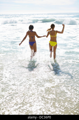 Friends in bikinis holding hands and running into ocean Stock Photo